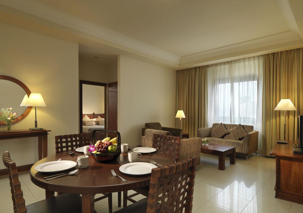Sunway Putra Hotel Kuala Lumpur Room photo The image shows an inviting and well-decorated room, likely a hotel suite or a cozy living area. In the foreground, there is a round dining table set for four, complete with plates and a centerpiece of fruit. Surrounding the table are wooden chairs w