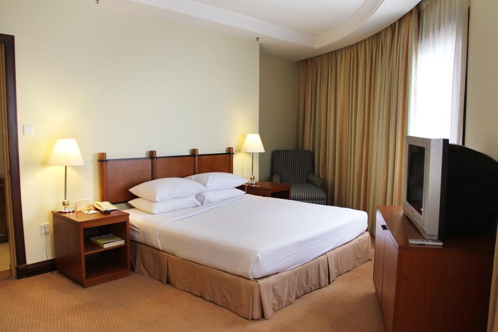 Sunway Putra Hotel Kuala Lumpur Room photo The photo shows a simple yet stylish hotel room. It features a double bed with white bedding, a bedside table on either side with lamps. There is a comfortable armchair in one corner and a television on a wooden cabinet. The room has large windows co